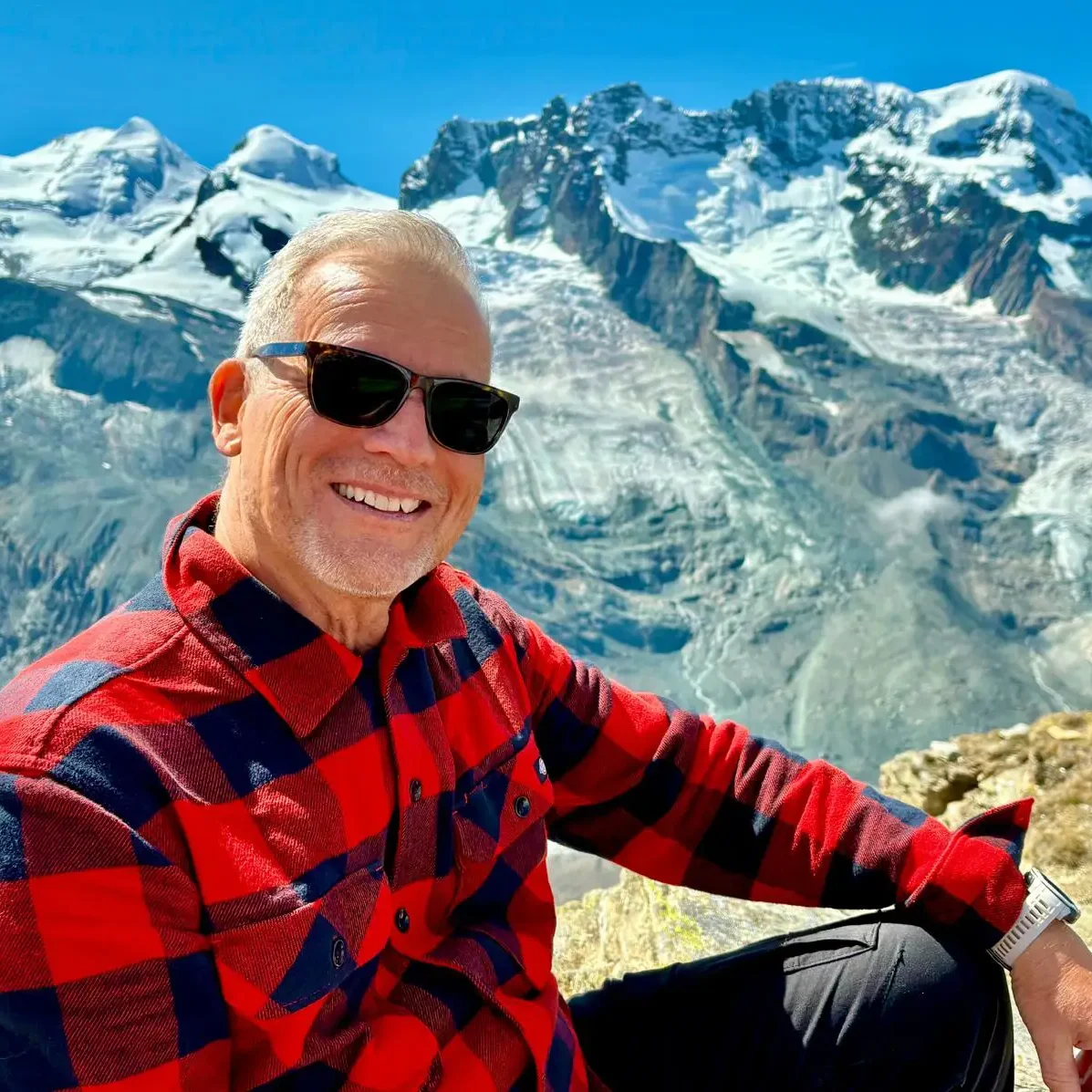Mann mit rotem kariertem Hemd und Sonnenbrille sitzt vor einer schneebedeckten Hochgebirgslandschaft mit Gletschern und blauem Himmel.