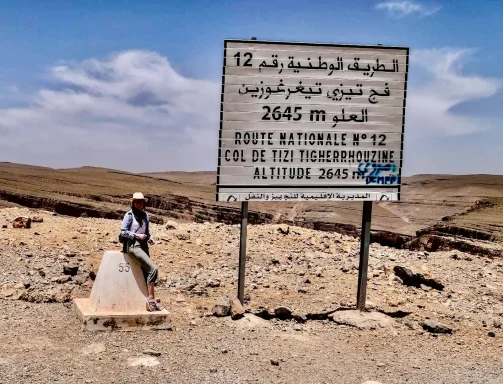 Frau am Gebirgspass Tizi n’Tichka in Marokko neben einem Straßenschild auf 2.645 Metern Höhe, umgeben von einer felsigen Gebirgslandschaft.