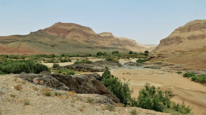 Weite Gebirgslandschaft in Marokko mit kargen, geschichteten Felsbergen, einem trockenen Flussbett und grüner Vegetation in einem Wüstental.