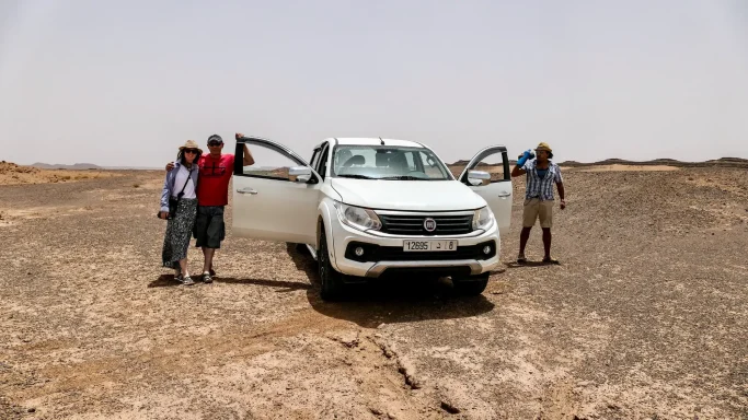 Drei Personen mit einem weißen Fiat-Pick-up in der kargen Wüstenlandschaft Marokkos bei einem Ausflug zu einer Fossilienfundstätte.